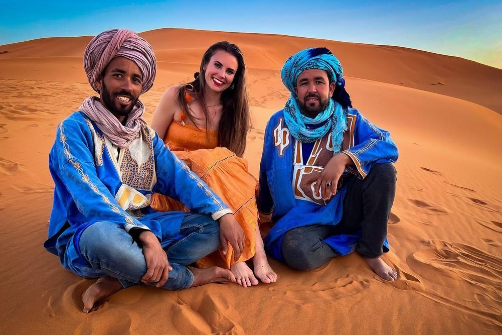 A smiling traveler sitting with local Berber guides Mohamed and Brahim in the Merzouga desert, showcasing authentic Morocco Grand Tours hospitality.